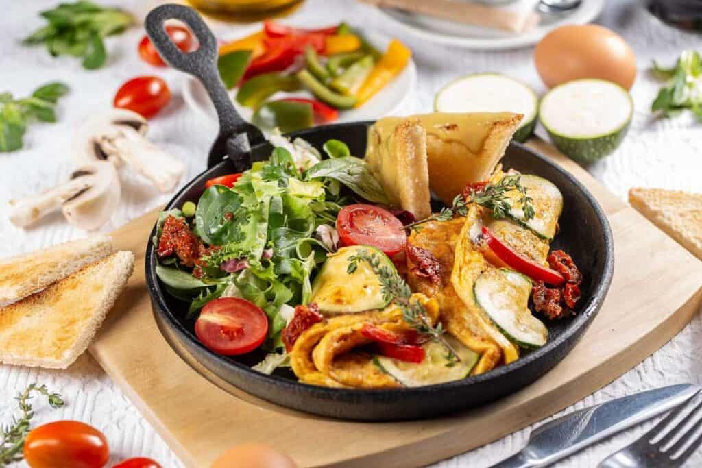 A skillet with an omelette, cherry tomatoes, salad, and a bread slice on a wooden board. Surrounding items include sliced mushrooms, bell peppers, eggs, and toast triangles—perfect for breakfast for dinner.