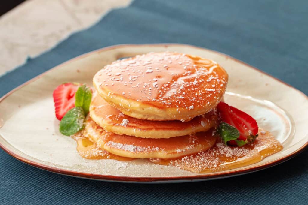 A stack of three pancakes drizzled with syrup and sprinkled with powdered sugar, garnished with sliced strawberries and mint leaves, rests on a white plate against a blue tablecloth—perfect for breakfast for dinner.