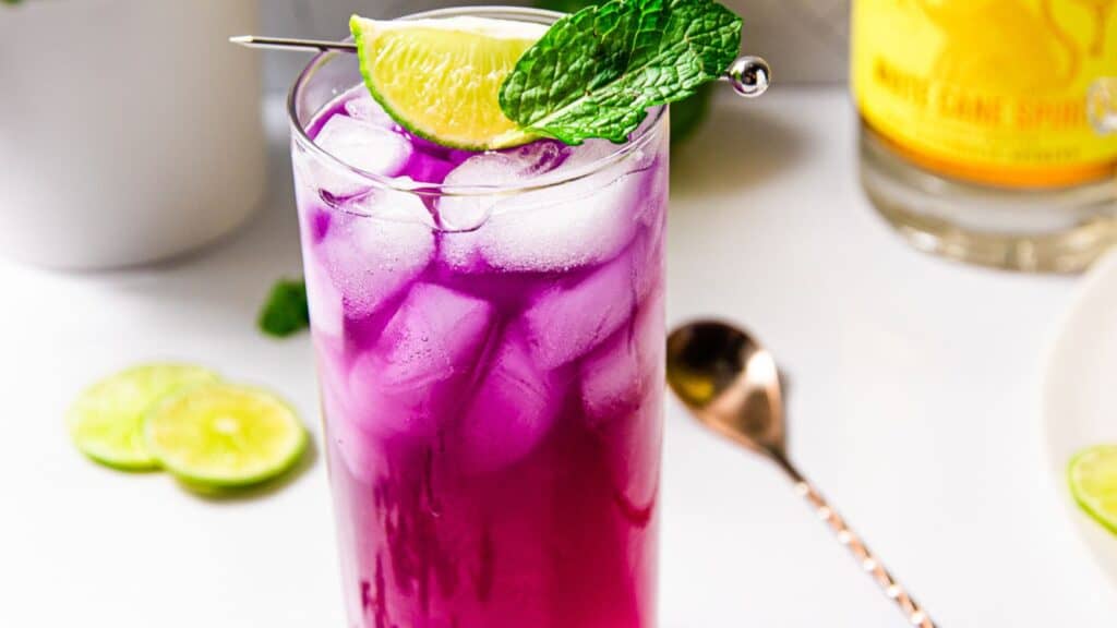 A vibrant purple cocktail with ice in a tall glass, garnished with a lime slice and mint leaf, beside a bottle and lime slices on a white surface.