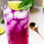 A vibrant purple cocktail with ice in a tall glass, garnished with a lime slice and mint leaf, beside a bottle and lime slices on a white surface.