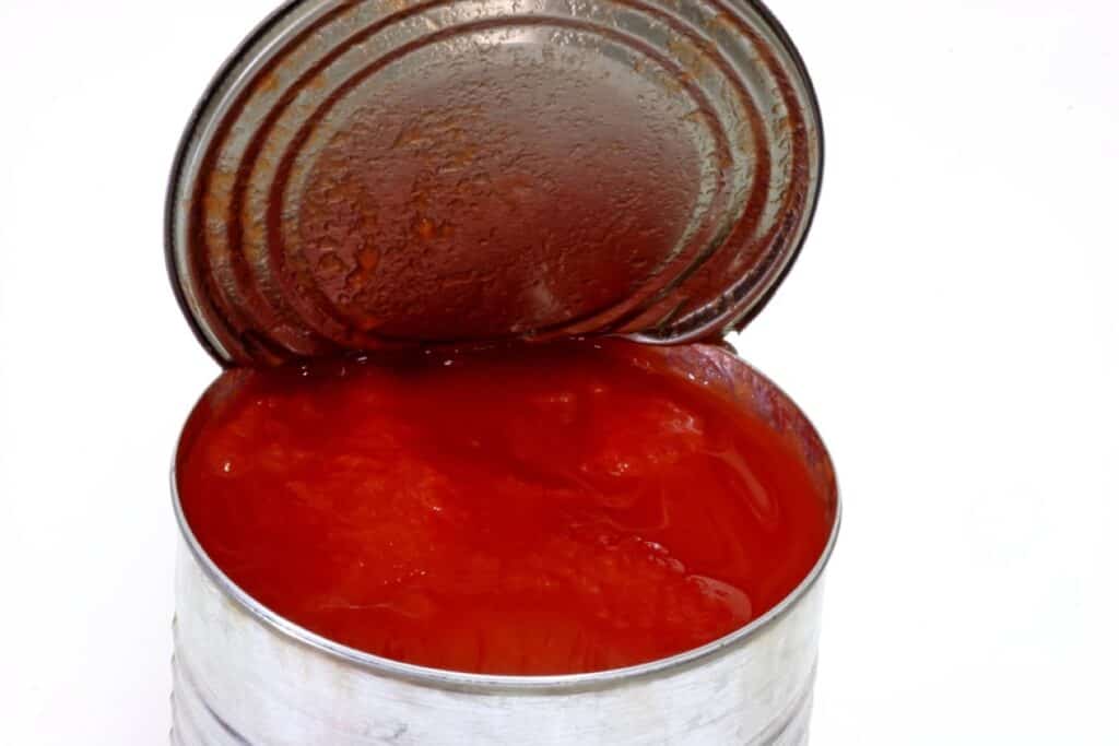 An open tin can with tomato sauce inside, showing the sauce's bright red color against the steel can and its partly peeled lid.