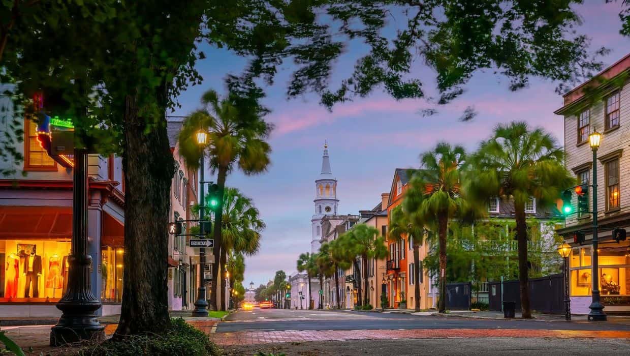 Dusk view of a tree-lined street with historic buildings and a church steeple, ideal for things to do in Charleston with kids, lit with streetlights under a colorful sky.