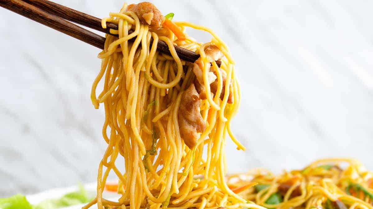 Chopsticks holding a chicken lo mein with noodles, vegetables, and chicken, set against a white background.