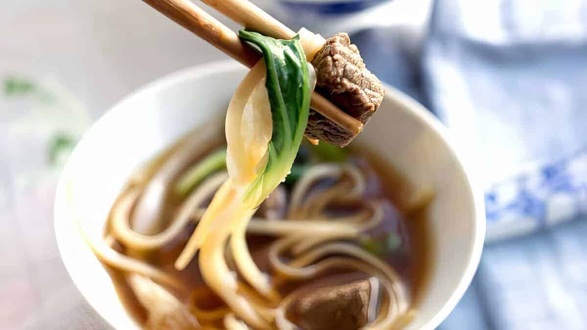 A white bowl containing beef noodle soup with a dark broth, slices of beef, white noodles, and green scallions on top.