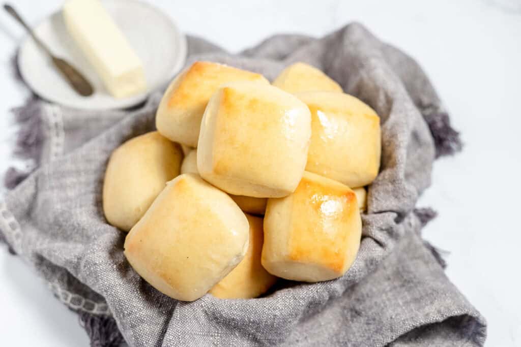 A pile of freshly baked dinner rolls on a cloth, with a stick of butter and a small dish in the background on a marble surface, resembling those from copycat pro recipes.