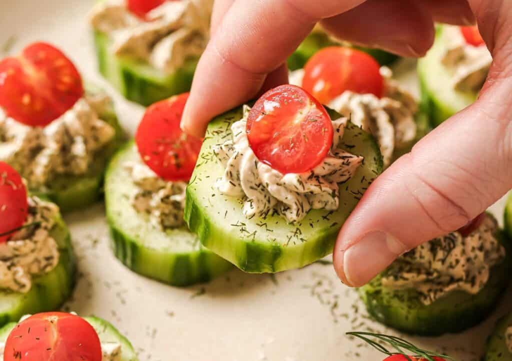 Hand holding a cucumber slice topped with herbed cream cheese and half a cherry tomato, surrounded by similar appetizers on a platter.