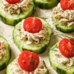 Sliced cucumbers topped with dollops of herb cream cheese, cherry tomato halves, and dill sprigs arranged on a beige surface.
