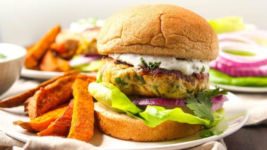 A grilled burger topped with lettuce, red onion, and a creamy white sauce is served with a side of sweet potato fries on a white plate.
