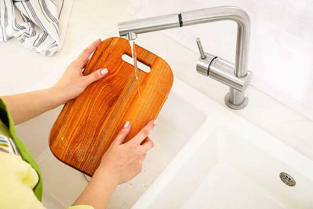 Hands carefully washing a wooden cutting board under a running faucet in a white sink.