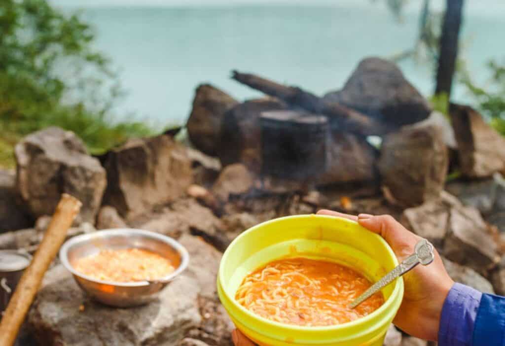 Person holding a yellow bowl of noodles with a campfire and cooking pot in the background near a lake.