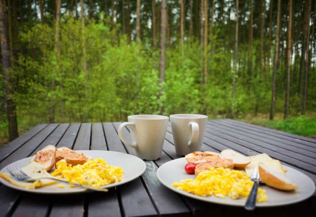 Two plates of scrambled eggs, bread, and tomatoes accompanied by two mugs on a wooden table outdoors with a forest backdrop.