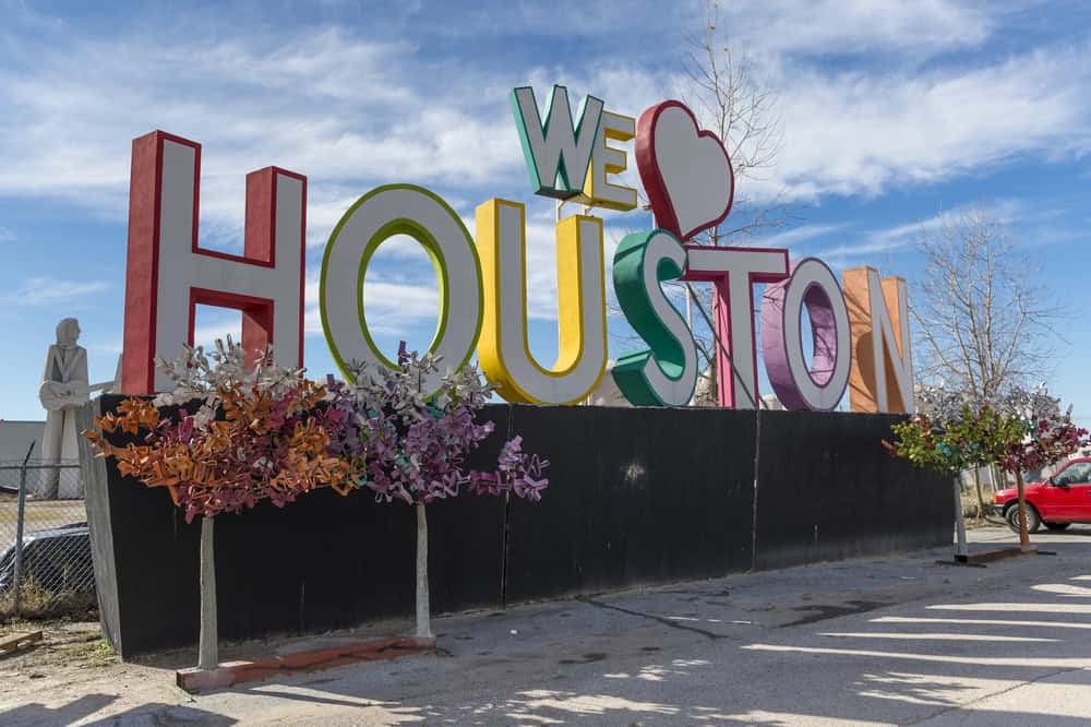 A colorful sign reads "We Heart Houston" with metal floral decorations in front, set against a blue sky.