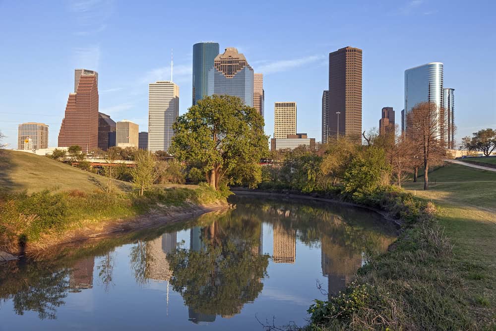 Houston skyline reflected in a tranquil river at sunset with clear skies and surrounding greenery.