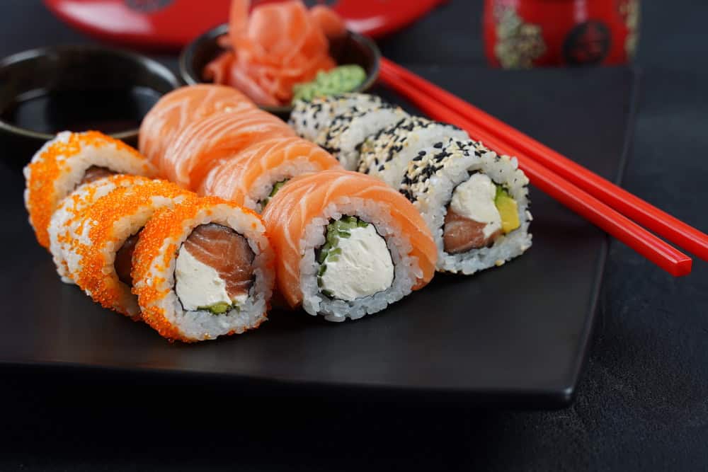 A black plate with three sushi rolls, topped with fish roe, fish slices, and sesame seeds. Ginger, wasabi, soy sauce, and red chopsticks are on the side.