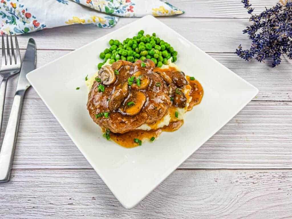 A plate of Salisbury steak with gravy on mashed potatoes, and a side of peas on a white square dish. The meal is set on a white wooden table beside silver cutlery and a floral napkin.