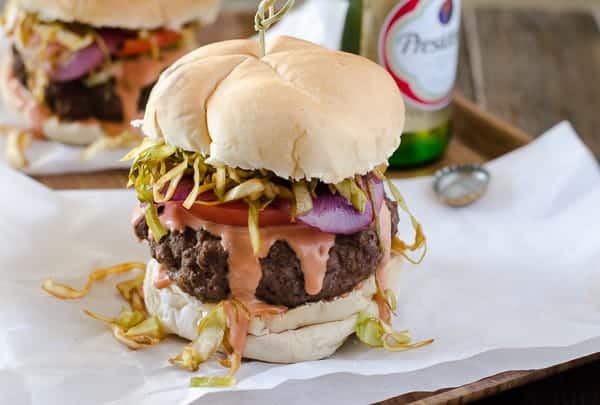 A hamburger with a beef patty, shredded lettuce, sliced tomato, onion, and sauce, topped with a bun. A bottle is visible in the background.