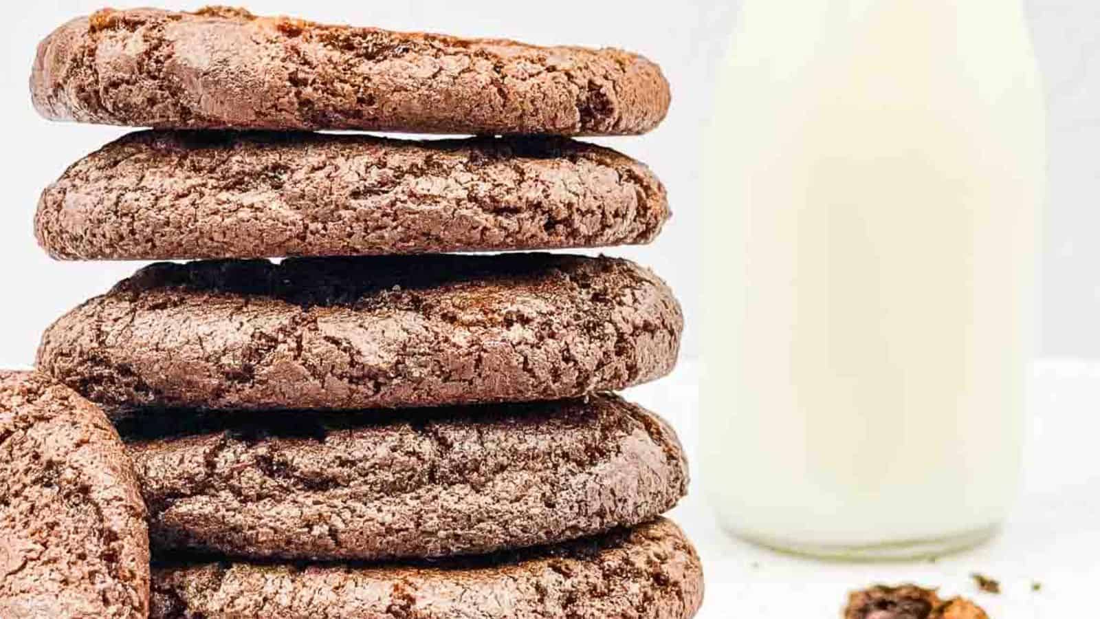 A stack of chocolate cookies with a bottle of milk in the background on a white surface.