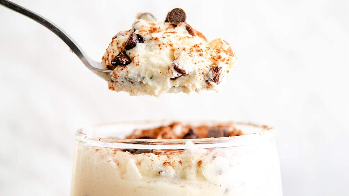 A spoon lifting a portion of creamy ricotta dessert sprinkled with chocolate chips and a dash of cinnamon, with more of the dessert visible in the glass below.