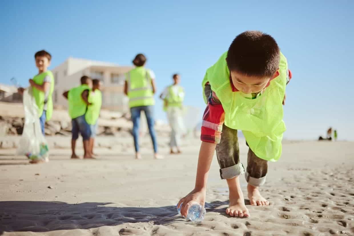 Children wearing neon yellow safety vests clean up a beach by collecting plastic bottles and other debris.