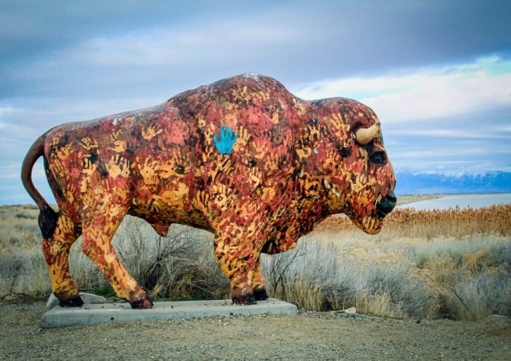 A bison statue decorated with colorful handprints stands on an island facing the water.