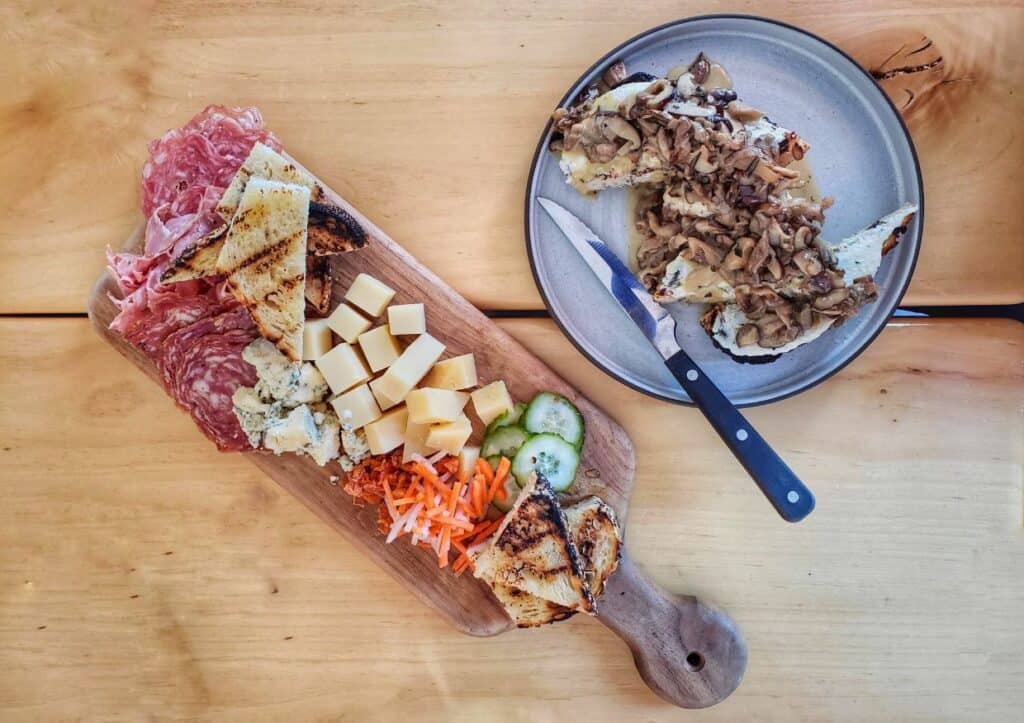 A wooden board with meats and cheeses sits on a wooden table next to a plate of mushroom crostini.