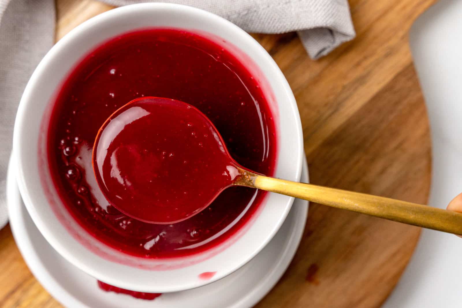 A bowl filled with red berry sauce. A golden spoon is partially submerged in the sauce, resting on the edge of the bowl.