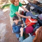 A family packs camping gear into the trunk of their car, with a happy child holding a fishing rod, and parents arranging bags.