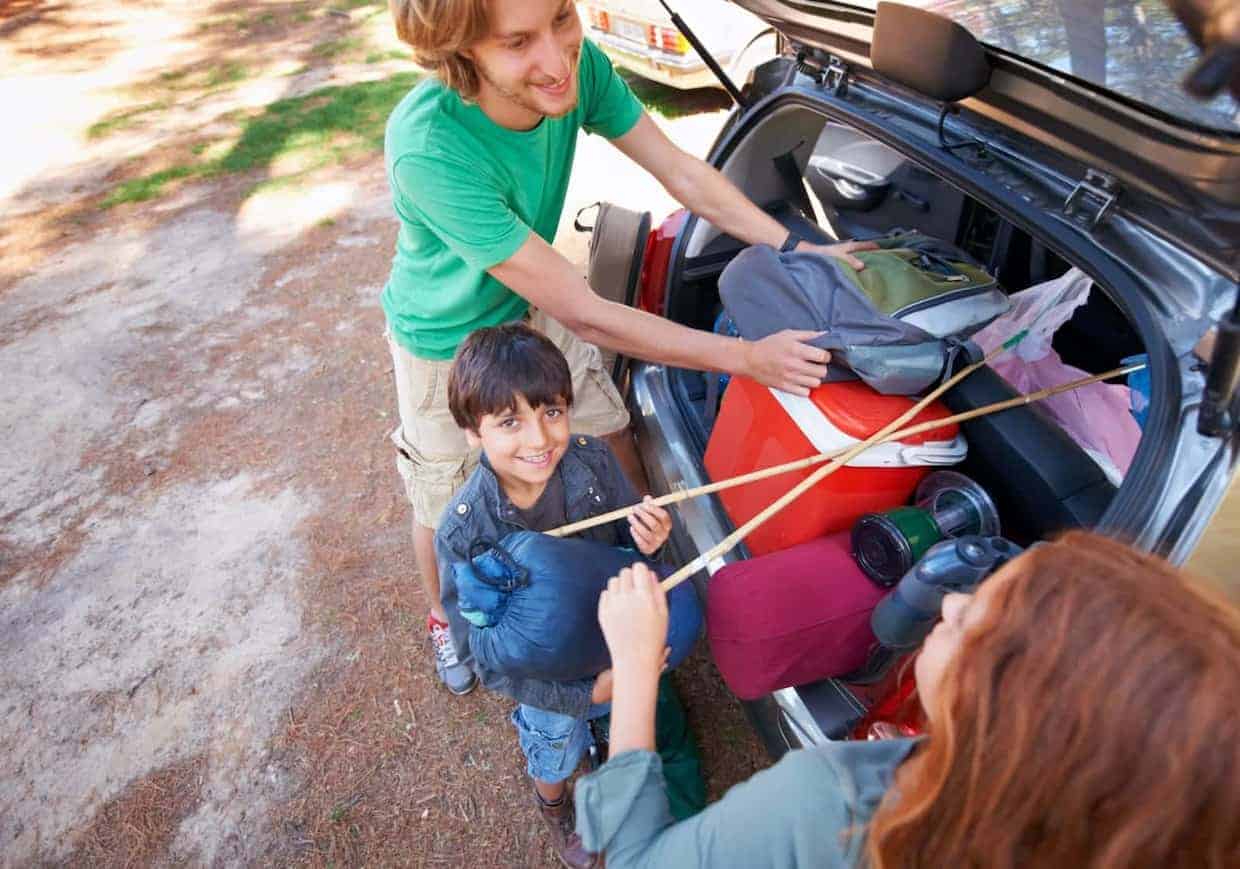 A family packs camping gear into the trunk of their car, with a happy child holding a fishing rod, and parents arranging bags.