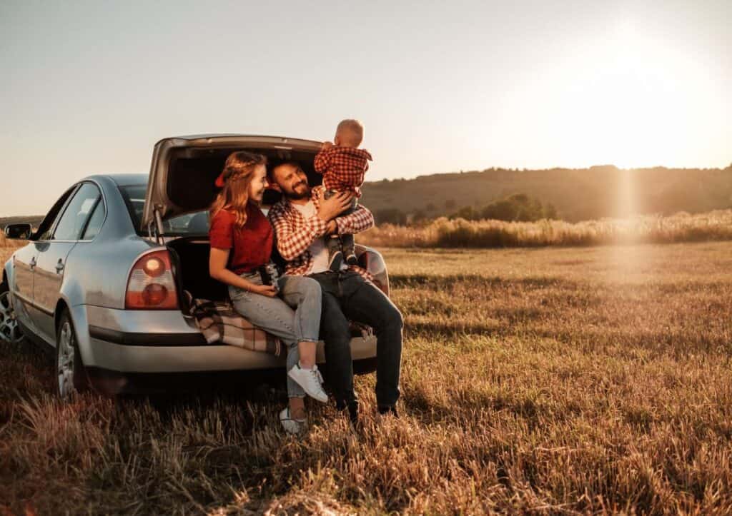 A family of three sits on the edge of an open car trunk in a field at sunset, with a scenic landscape in the background.