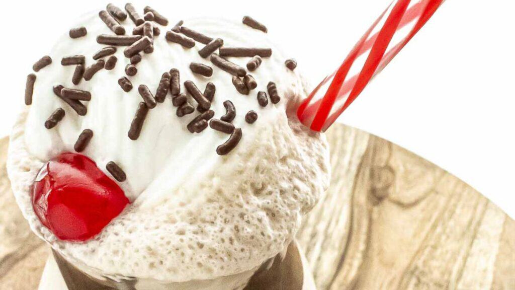 Close-up of a milkshake topped with whipped cream, chocolate sprinkles, and a cherry, served in a glass with a red striped straw.