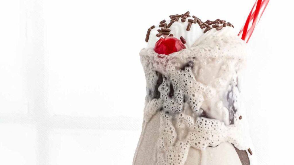 A close-up of a melting milkshake with a cherry on top and chocolate sprinkles, served with a red straw, against a white background.