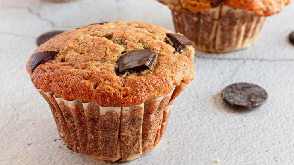 Close-up of a chocolate chip muffin on a light background, with more muffins blurred in the background.