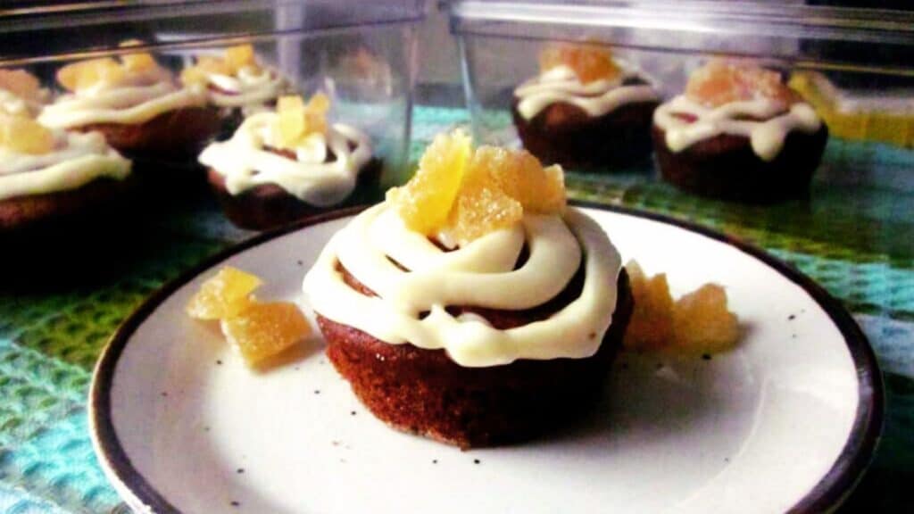 A plate with a frosted cupcake topped with candied ginger, surrounded by similar cupcakes in clear containers, perfect for successful brunch hosts.