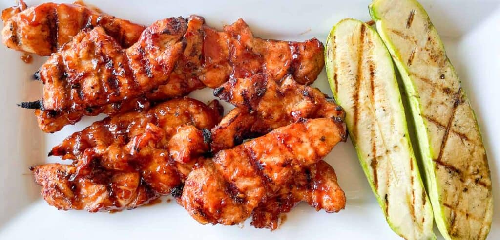 Grilled barbecue chicken pieces and grilled zucchini slices on a white plate.