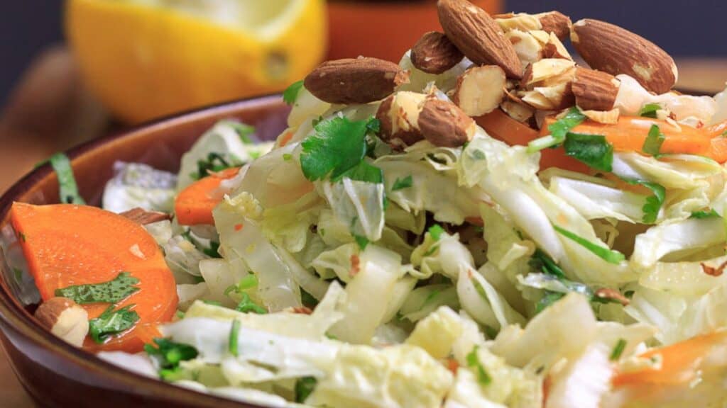 A bowl of fresh salad containing lettuce, carrots, chopped almonds, and herbs with a halved lemon in the background.