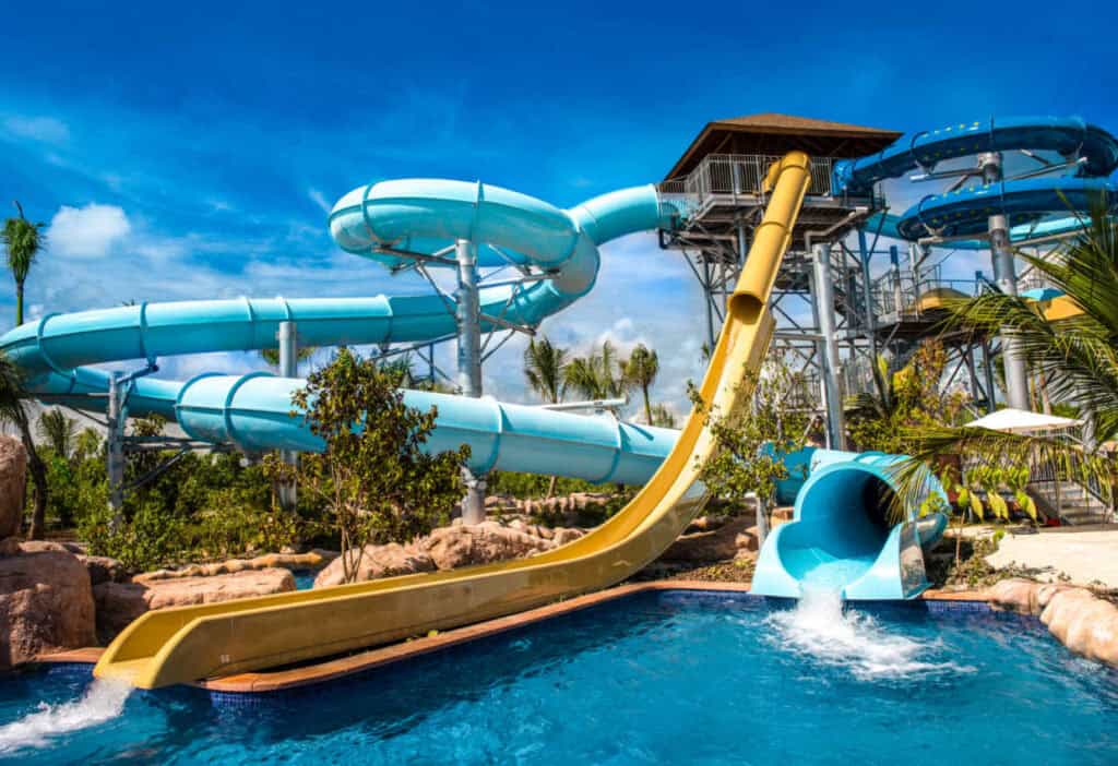 A water park with multiple water slides, including a yellow slide and a blue tube slide, ending in a pool on a sunny day with clear skies.