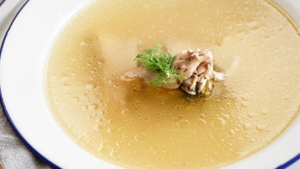 A bowl of clear soup with chicken pieces and a sprig of dill in the center, served in a white bowl with a blue rim.