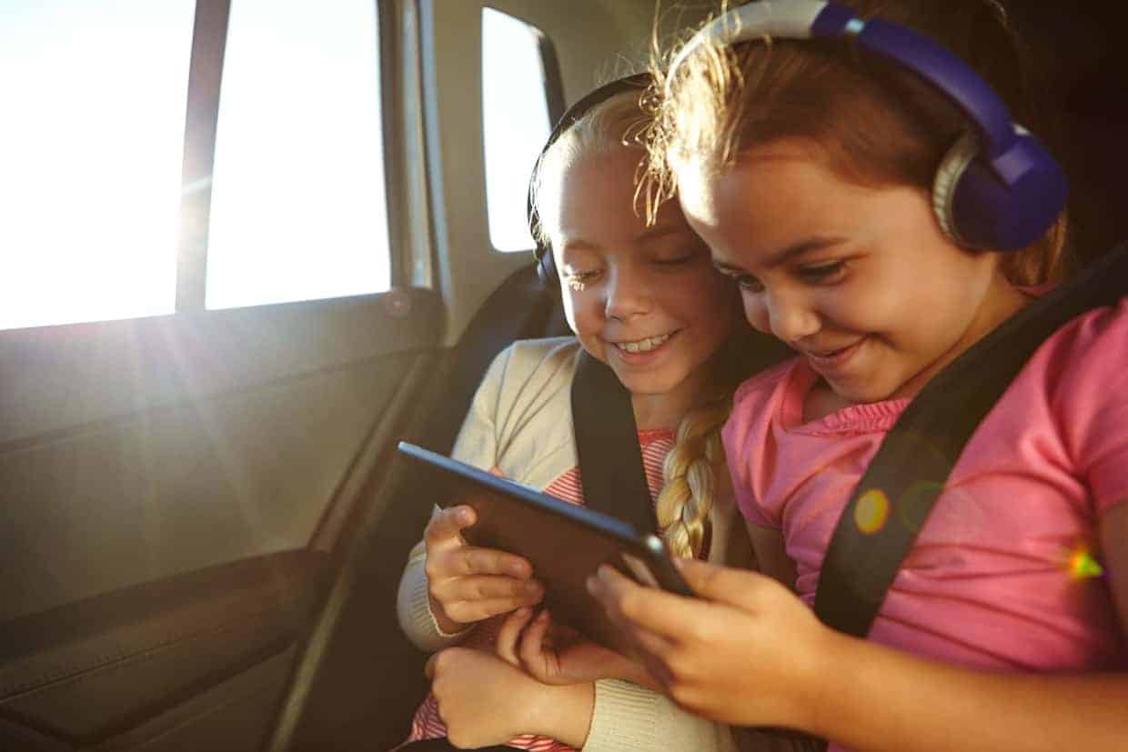 Two young girls with headphones smiling and looking at a tablet together inside a car on a family road trip with kids.