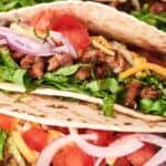 Close-up of three soft tacos filled with seasoned ground meat, shredded lettuce, diced tomatoes, grated cheese, and sliced red onions.