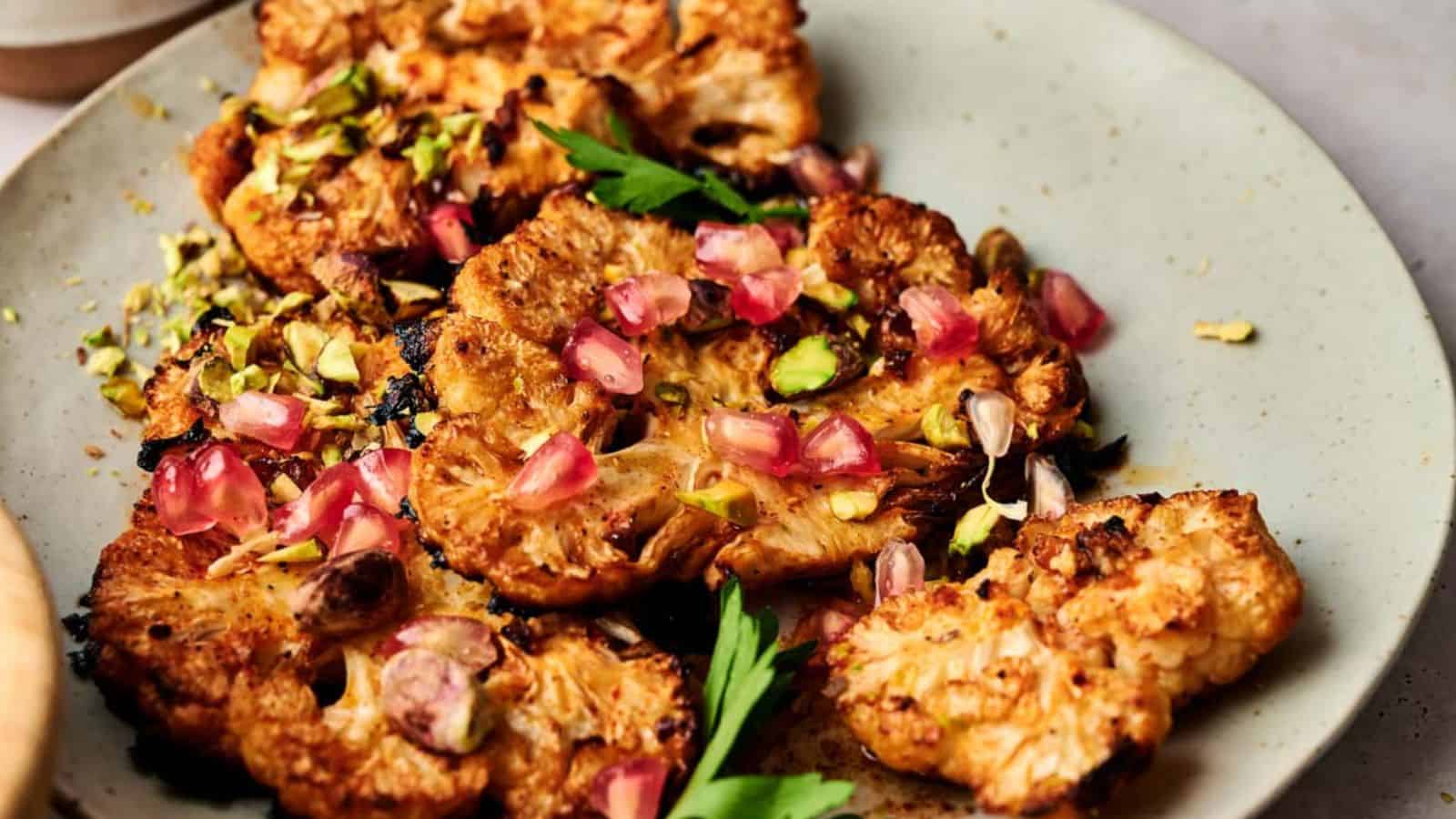 A plate of roasted cauliflower steaks garnished with chopped pistachios, pomegranate seeds, and fresh parsley.