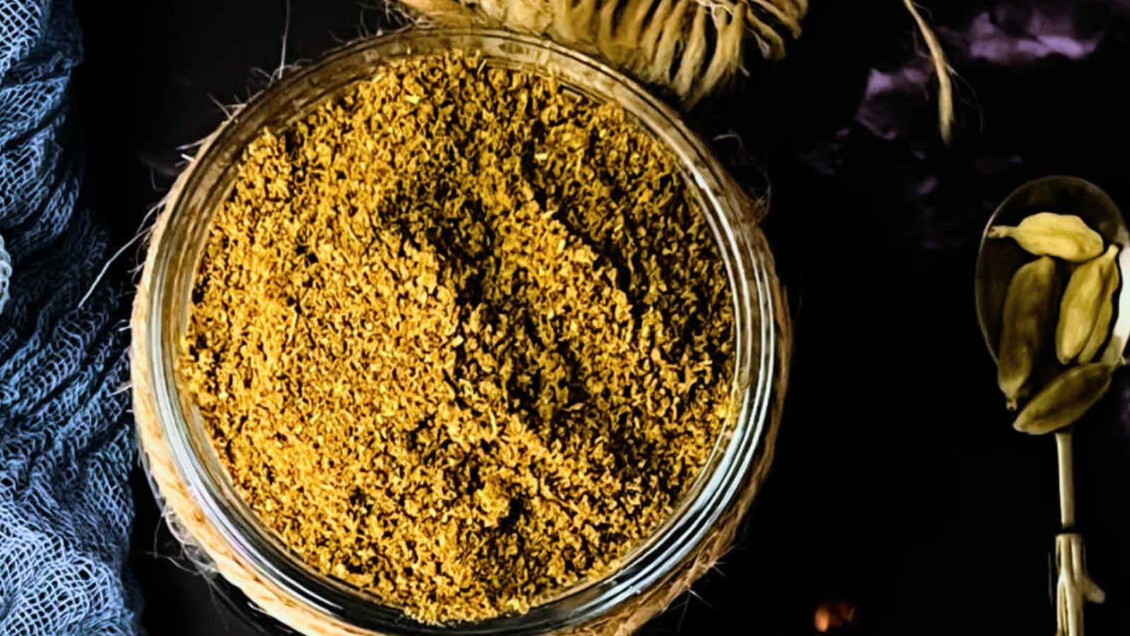 A glass jar filled with ground spices seen from above, placed on a dark surface with some whole cardamom pods in the background.