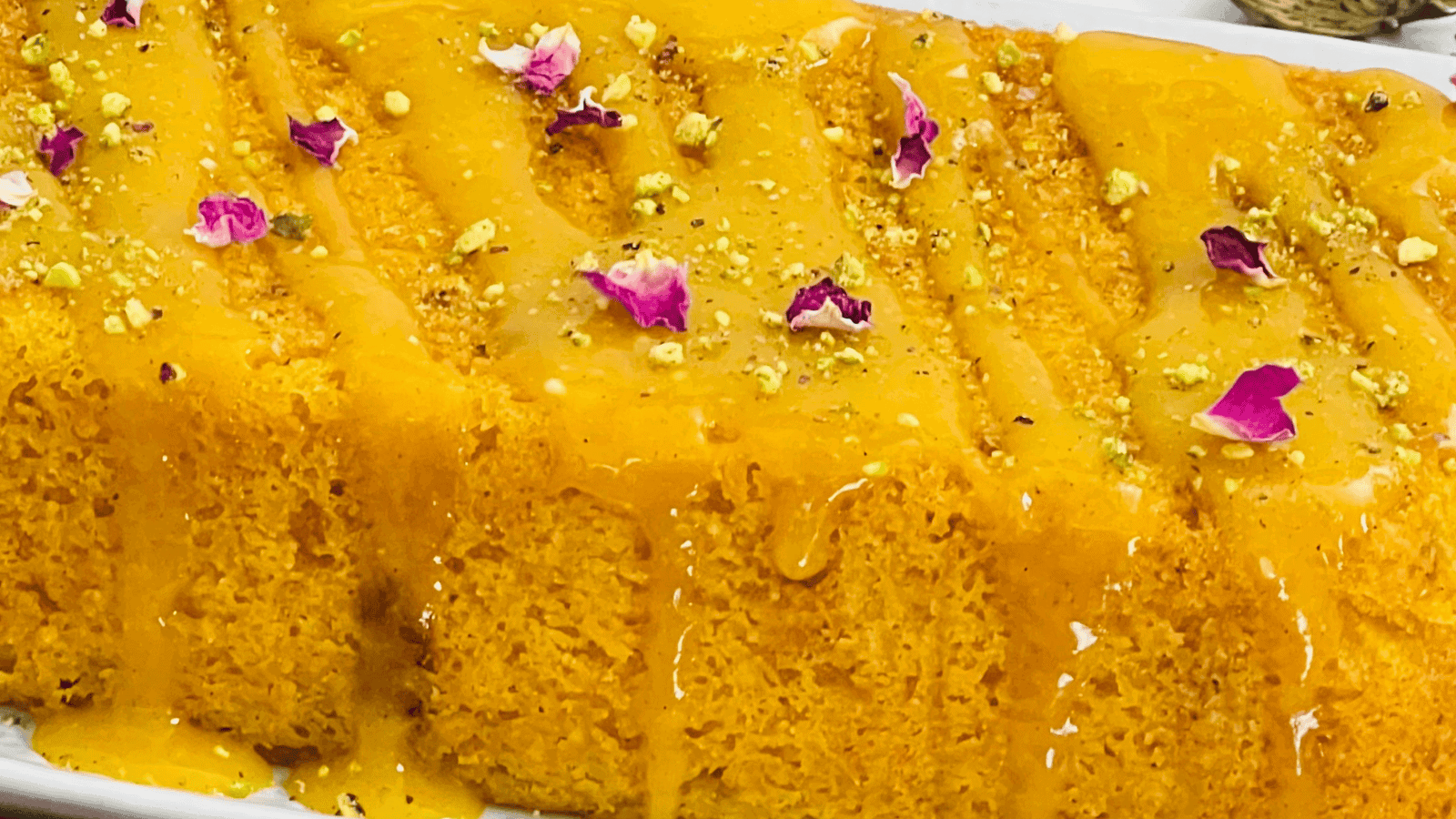 Close-up of a vibrant orange dessert garnished with flower petals and nuts, drizzled with syrup on a white rectangular plate.