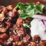 Close-up of a bowl of chili, topped with chopped cilantro, sliced red onions, and a dollop of sour cream. The chili contains beans, tomatoes, and other visible vegetables.