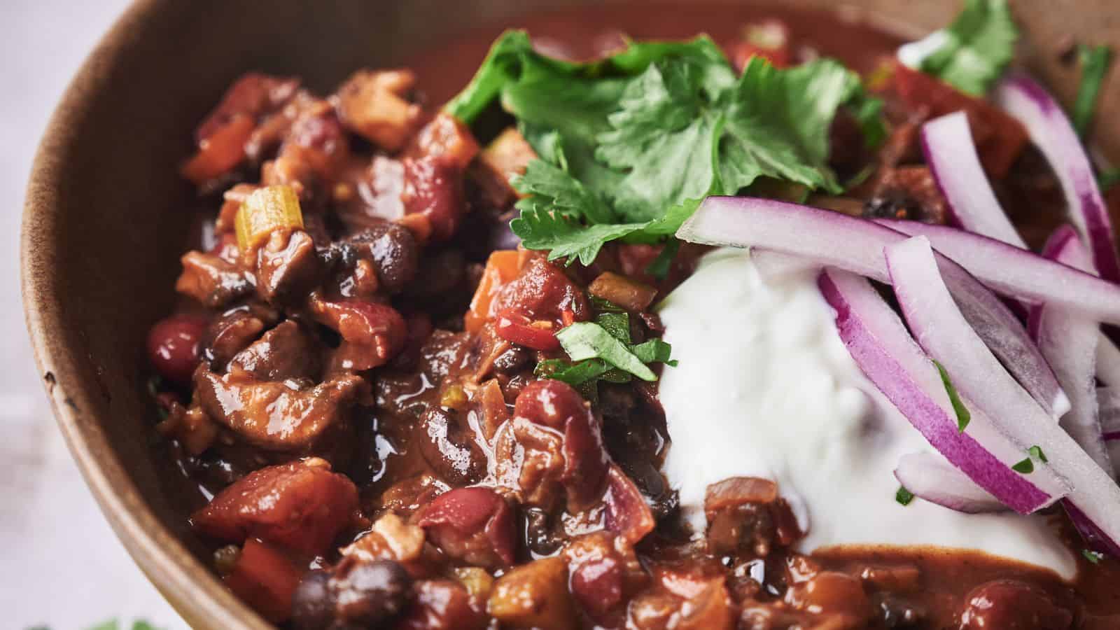 Close-up of a bowl of chili, topped with chopped cilantro, sliced red onions, and a dollop of sour cream. The chili contains beans, tomatoes, and other visible vegetables.