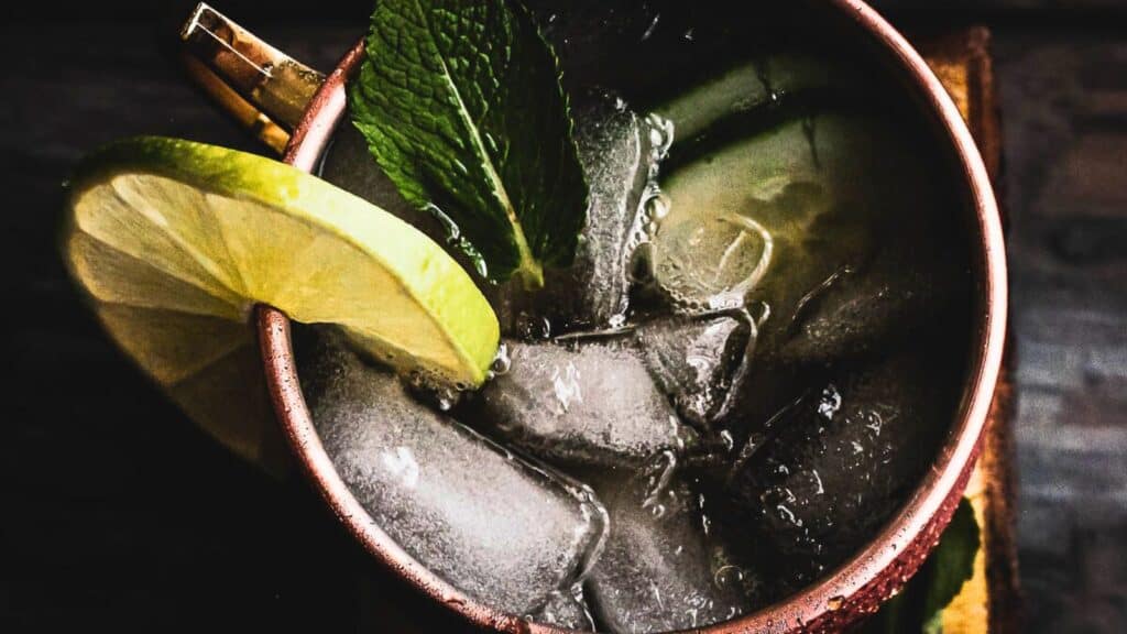 Close-up of a moscow mule cocktail in a copper mug with ice, garnished with a lime wedge and fresh mint on a dark wooden surface.