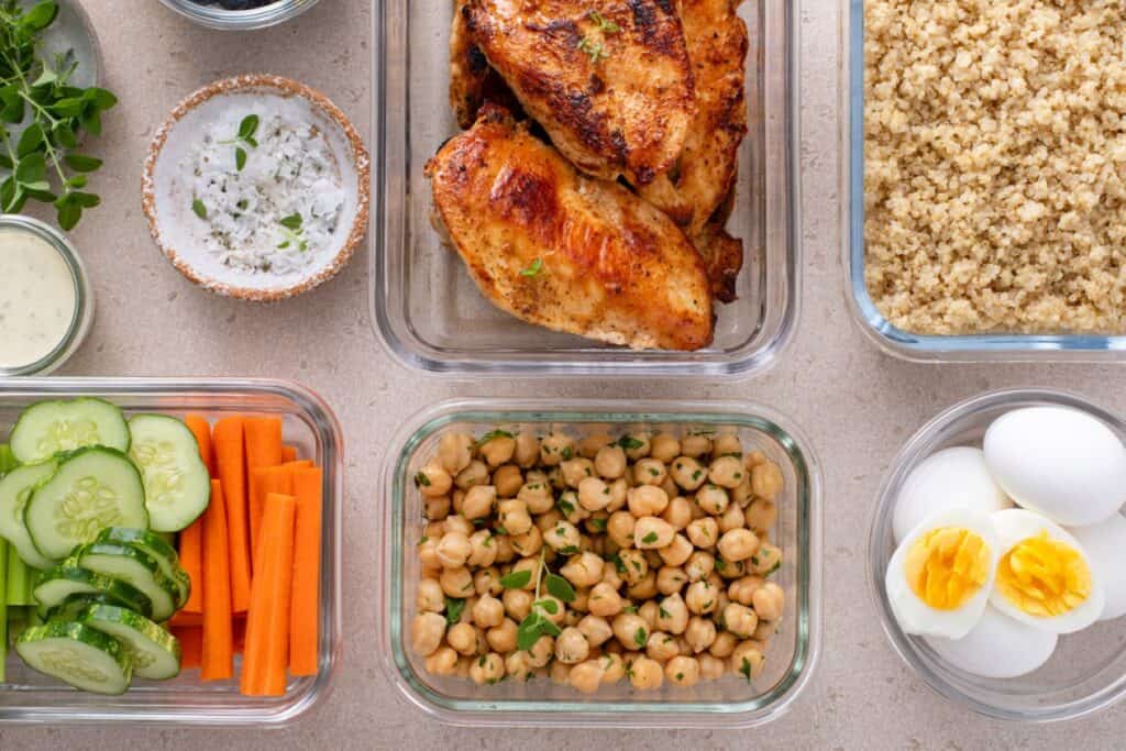 Top view of a meal prep setup with grilled chicken, quinoa, chickpeas, boiled eggs, cucumber and carrot sticks, salt, black pepper, and a small bowl with dressing.