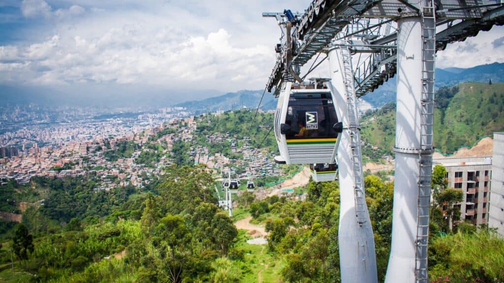 A cable car travels above a lush green hillside with a panoramic view of a densely populated city valley below, under a cloudy sky.