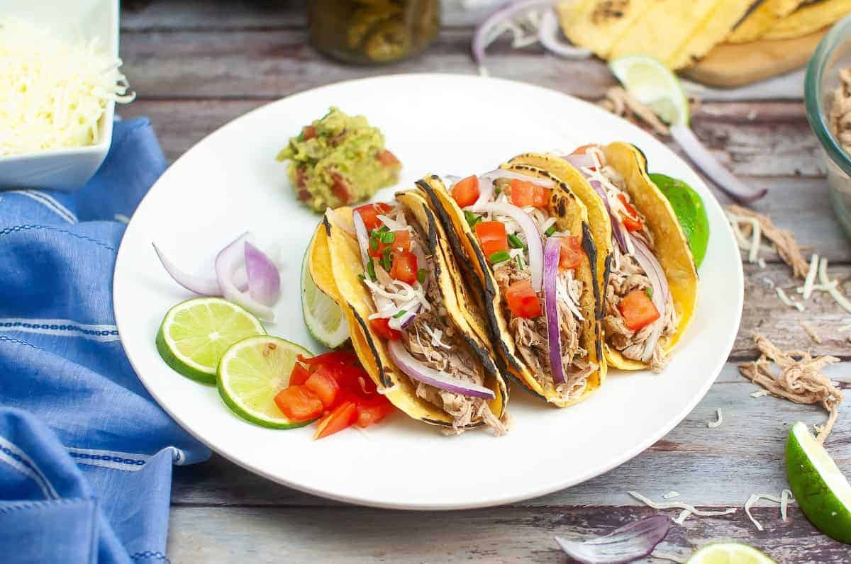 Pork carnitas in taco shells with lime, guacamole, onion, and tomato on white plate on wood table.