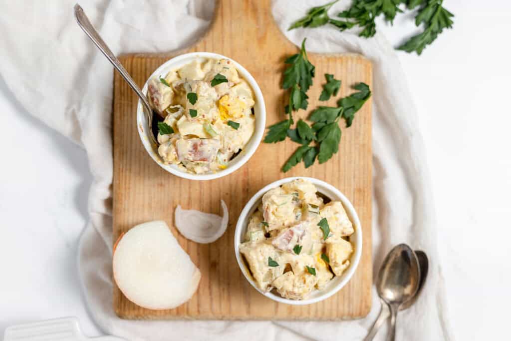 Two small white bowls filled with potato salad are placed on a wooden cutting board. Fresh parsley, a half onion, and two spoons are also on the board. A white napkin is draped underneath—perfect for summer cookout salads.