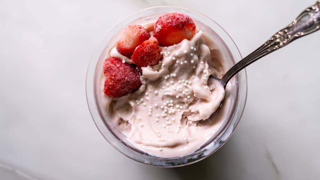 A bowl of strawberry ice cream topped with frozen strawberries and white sprinkles, with a silver spoon inserted into it, placed on a white surface.